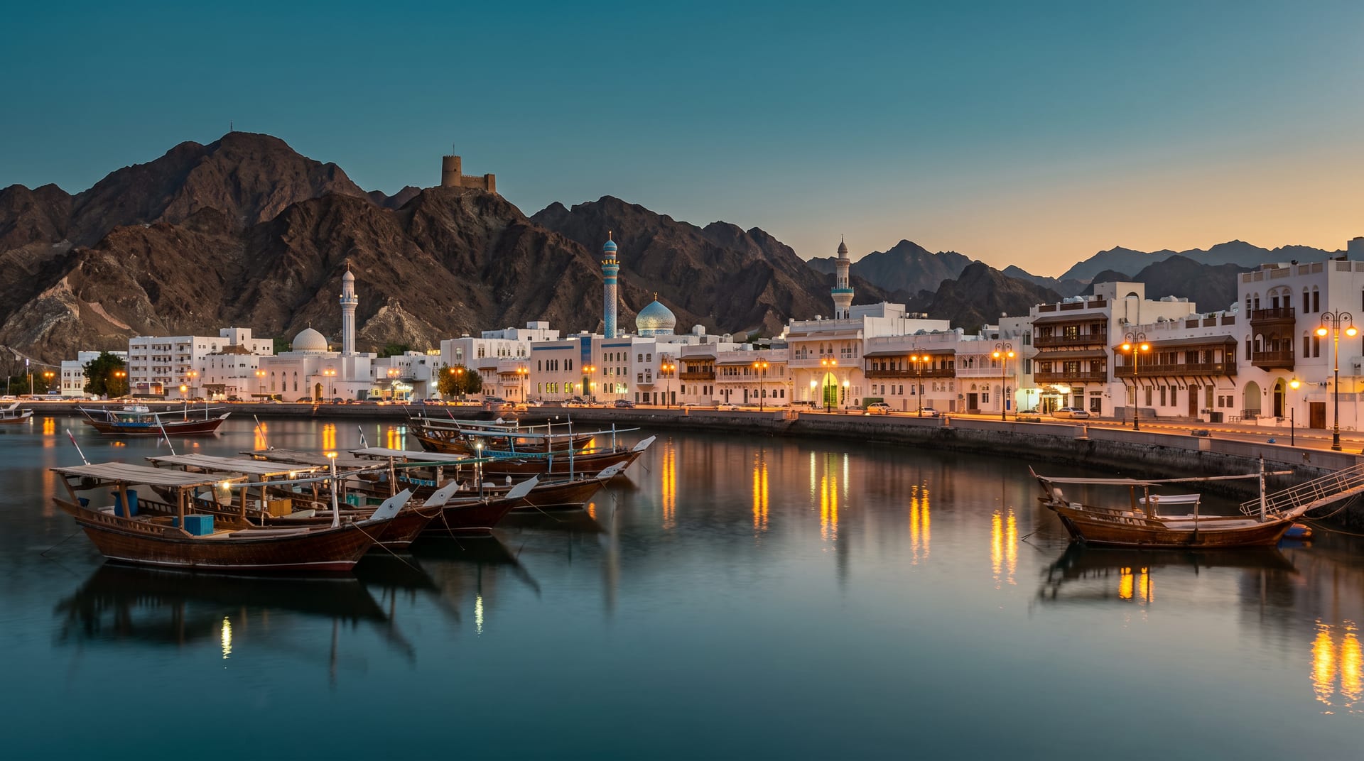 The Mutrah Corniche in Muscat at dusk, with dhows in the harbour and the Hajar mountains behind